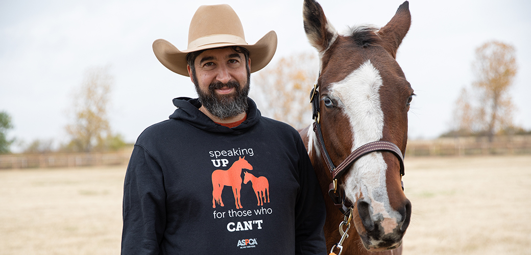 A man with a cowboy hat, salt and pepper beard, and an aspca horse hoodie next to a brown and white horse